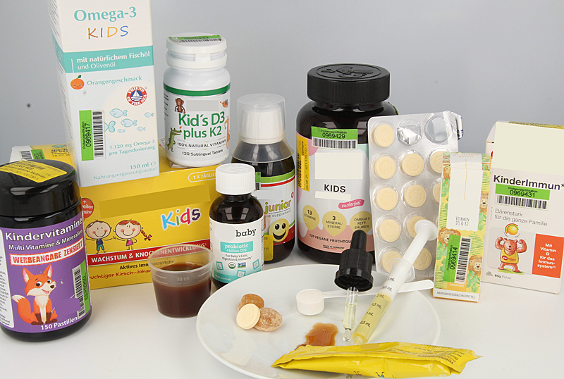 Zu sehen sind verschiedene Nahrungsergänzungsmittel, die mit bunten Aufmachungen und niedlichen Grafiken speziell auf die Zielgruppe Kinder abzielen. Zu sehen sind auch die verschiedenen Darreichungsformen, darunter Lutschtabletten, Fruchtgummis, Pulver in einem Dosierlöffel, eine Tropfpipette sowie Flüssigkeiten in einem Portionsstick, einem Dosierbecher und einer Dosierspritze.